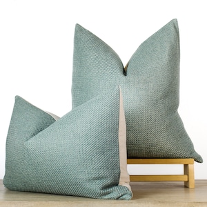 May include: Two teal throw pillows with a textured weave pattern. The pillows are displayed on a small wooden stool against a white background. The back of the pillows are a light beige color.