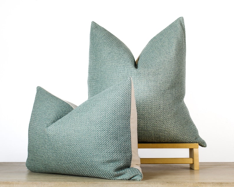 May include: Two teal throw pillows with a textured weave pattern. The pillows are displayed on a small wooden stool against a white background. The back of the pillows are a light beige color.