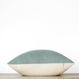 May include: A decorative pillow with a two-tone design. The top portion is a textured teal fabric, while the bottom is a cream-colored canvas. The pillow is placed on a wooden surface against a white background. The pillow is designed for home decor.