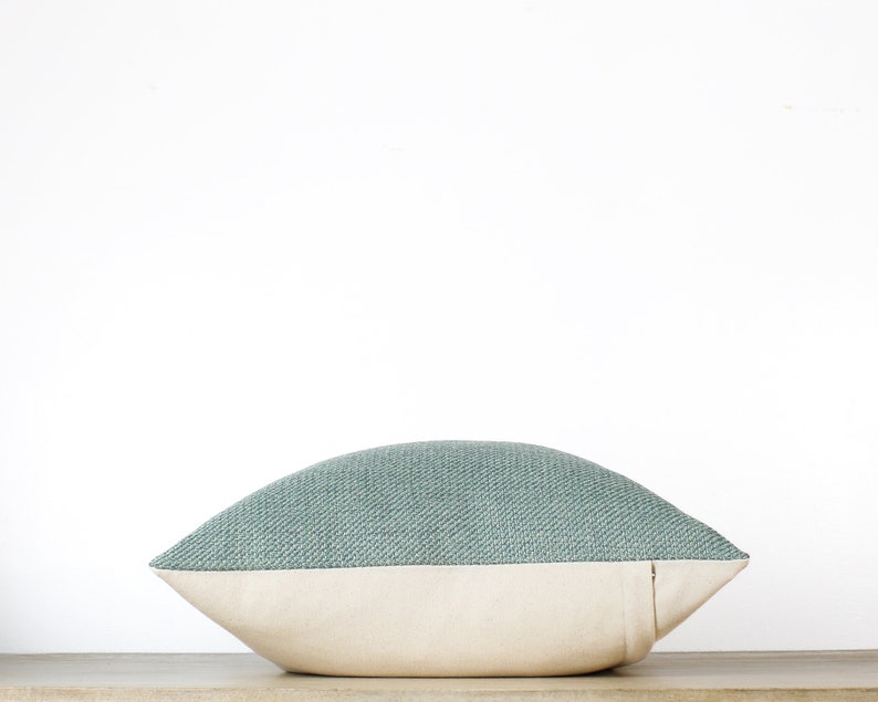 May include: A decorative pillow with a two-tone design. The top portion is a textured teal fabric, while the bottom is a cream-colored canvas. The pillow is placed on a wooden surface against a white background. The pillow is designed for home decor.