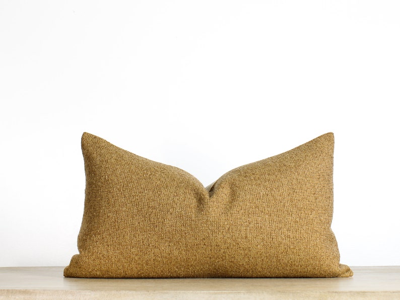 May include: A rectangular decorative pillow in a warm, golden-brown color. The pillow has a textured, woven appearance and is placed against a white background. The pillow is approximately 24 inches long.