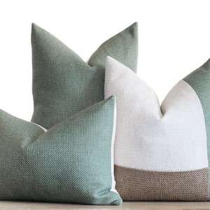 May include: Three decorative throw pillows in shades of green, white, and brown. The pillows have textured fabric and a simple, modern design. The pillows are arranged on a light-colored surface, with a white background.