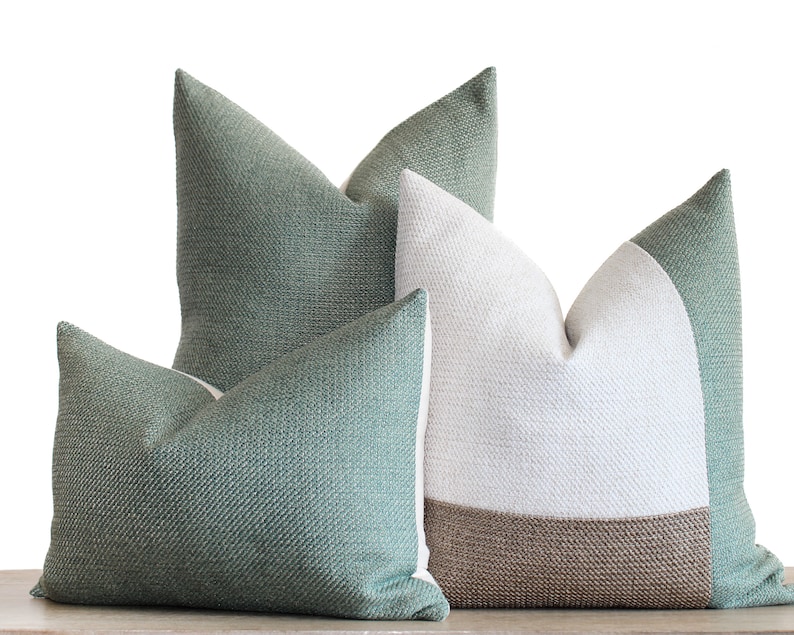 May include: Three decorative throw pillows in shades of green, white, and brown. The pillows have textured fabric and a simple, modern design. The pillows are arranged on a light-colored surface, with a white background.