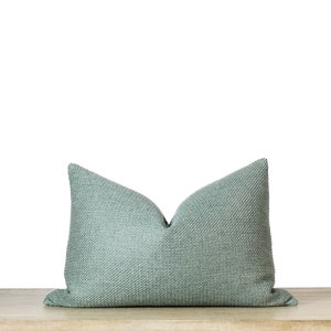 May include: A rectangular, light teal pillow with a textured weave pattern. The pillow is resting on a light-colored wooden surface against a white background. The pillow's shape is a classic bolster style.