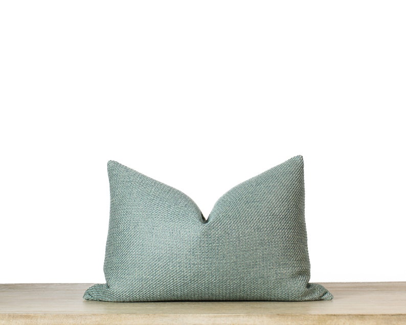 May include: A rectangular, light teal pillow with a textured weave pattern. The pillow is resting on a light-colored wooden surface against a white background. The pillow's shape is a classic bolster style.