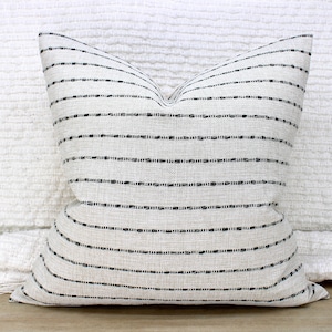 May include: Square decorative pillow with a neutral beige fabric and horizontal black stripes. The pillow has a textured appearance and a soft, inviting look. The pillow is a home decor item.