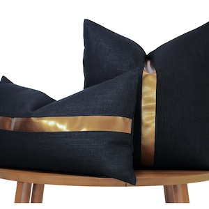 May include: Two dark charcoal black throw pillows with a textured fabric finish. One pillow is square and features a vertical metallic gold stripe down the center, while the other is rectangular and features a horizontal metallic gold stripe.