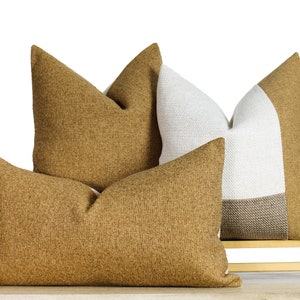 May include: Four decorative throw pillows in shades of tan and white. Two pillows are solid tan, one is white and tan, and one is a rectangular tan pillow. The pillows are arranged on a small wooden bench.