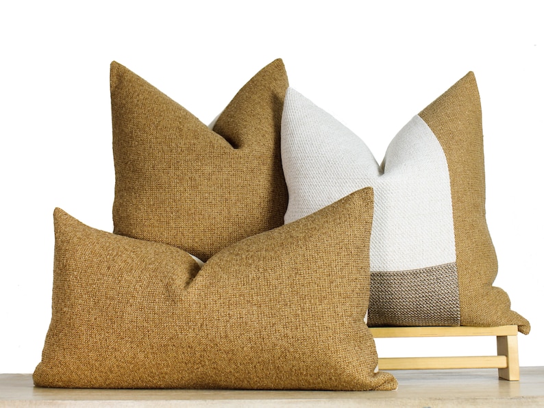 May include: Four decorative throw pillows in shades of tan and white. Two pillows are solid tan, one is white and tan, and one is a rectangular tan pillow. The pillows are arranged on a small wooden bench.