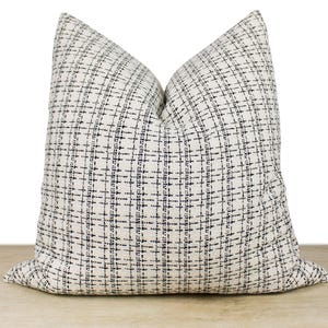 May include: A square decorative pillow with a cream-colored background and a navy blue grid pattern. The pillow is made of a textured fabric and is placed on a wooden surface.
