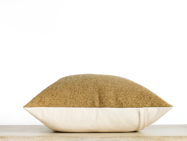 May include: A decorative pillow with a two-tone design. The top half features a textured, golden-brown fabric, while the bottom half is a smooth, off-white material. The pillow is rectangular and rests on a light-colored surface.