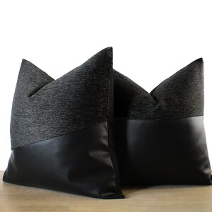 May include: Two decorative throw pillows. The top portion of each pillow is a dark gray woven fabric, while the bottom is black faux leather. The pillows are placed on a light brown surface against a white background.