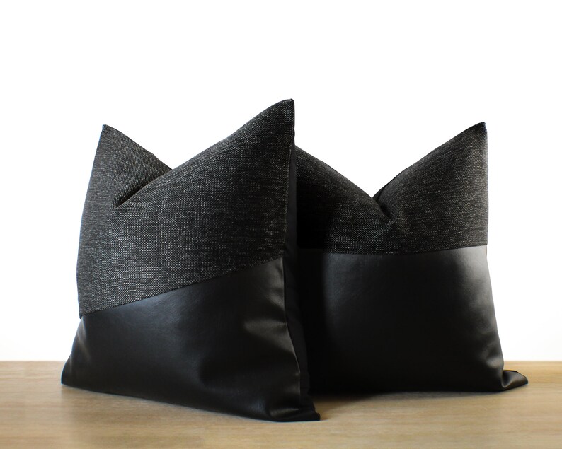 May include: Two decorative throw pillows. The top portion of each pillow is a dark gray woven fabric, while the bottom is black faux leather. The pillows are placed on a light brown surface against a white background.