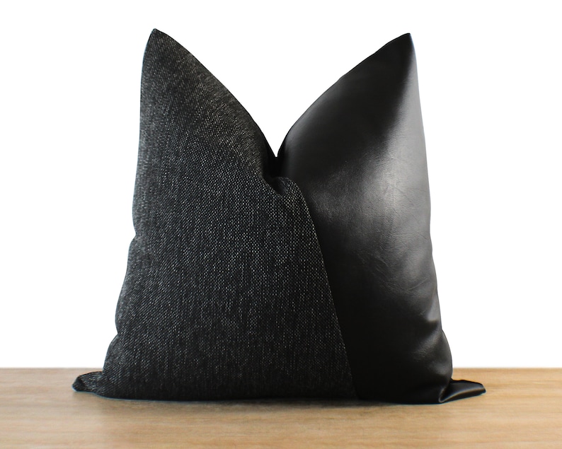May include: A decorative pillow with a two-tone design. One side features a dark gray textured fabric, while the other side is black faux leather. The pillow is rectangular and rests on a wooden surface.