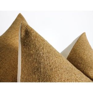 May include: Three decorative throw pillows with textured, golden-brown fabric. The pillows are arranged with their pointed tops facing upward, showcasing the neutral-colored, off-white fabric on the sides. The pillows are set against a plain white background.