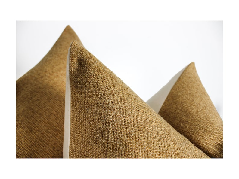 May include: Three decorative throw pillows with textured, golden-brown fabric. The pillows are arranged with their pointed tops facing upward, showcasing the neutral-colored, off-white fabric on the sides. The pillows are set against a plain white background.