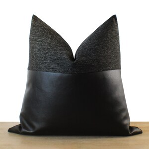 May include: A decorative pillow with a two-tone design. The top portion is a dark gray woven fabric, and the bottom is black faux leather. The pillow is square-shaped and has a soft, inviting appearance, suitable for home decor.