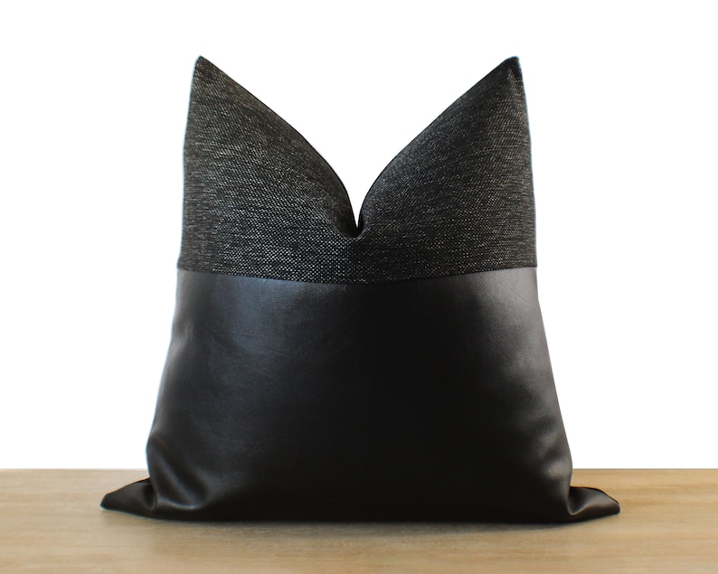 May include: A decorative pillow with a two-tone design. The top portion is a dark gray woven fabric, and the bottom is black faux leather. The pillow is square-shaped and has a soft, inviting appearance, suitable for home decor.