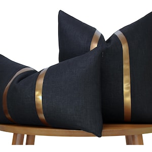 May include: Two black rectangular throw pillows featuring a vertical metallic bronze stripe running down the center of each cushion. The fabric has a visible woven texture, and the pillows are arranged with one positioned horizontally in front.