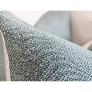 May include: Close-up of a teal-colored pillow with a textured weave. The pillow has a light-colored border and a soft, inviting appearance. The fabric has a subtle pattern, suggesting a comfortable home decor item.