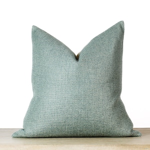 May include: A square, light teal pillow with a textured weave. The pillow is placed on a light-colored surface against a white background. The pillow has a classic, simple design, suitable for home decor.