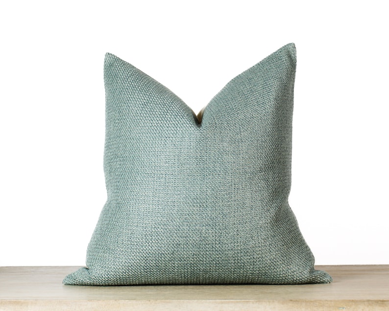 May include: A square, light teal pillow with a textured weave. The pillow is placed on a light-colored surface against a white background. The pillow has a classic, simple design, suitable for home decor.