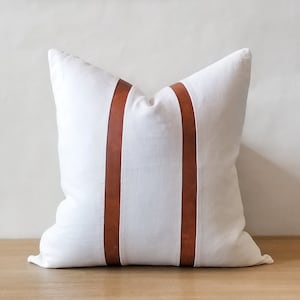 Può includere: Cuscino quadrato bianco con due fasce verticali in pelle marrone. Il cuscino ha un aspetto morbido e strutturato, con le fasce in pelle che offrono un dettaglio a contrasto. Il design è semplice e moderno, adatto all'arredamento.