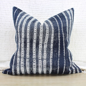 May include: A square decorative pillow with a navy blue and white striped pattern. The pillow has a textured appearance and is placed on a light-colored surface. The pillow's design features vertical stripes with dotted accents.