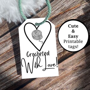 May include: A white heart-shaped tag with a black outline and a black and white illustration of a ball of yarn inside the heart. The tag has the text "Crocheted With Love!" printed on it. The tag is tied with a green string and is sitting on a wooden surface.  Cute & Easy Printable tags!