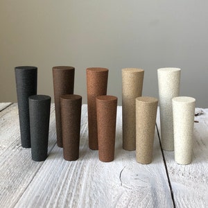 May include: A set of ten cylindrical ceramic bottle stoppers in various shades of brown, beige, and white. The stoppers are arranged in a row on a light wood surface.