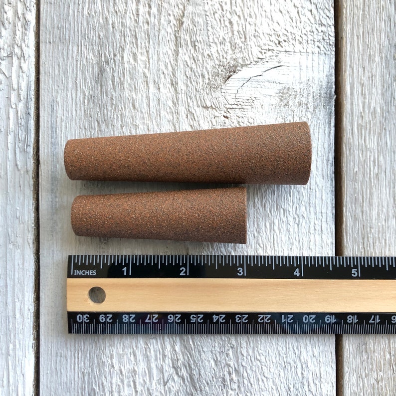 Puede incluir: Dos conos de lija marrones, uno ligeramente m&aacute;s grande que el otro, se muestran sobre una superficie de madera blanca. Una regla se coloca debajo de los conos, con los n&uacute;meros 1, 2, 3, 4 y 5 visibles.