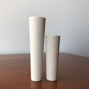 Puede incluir: Dos cilindros de cer&aacute;mica blanca, uno m&aacute;s alto que el otro, con una superficie texturizada. Los cilindros est&aacute;n sobre una superficie de madera.