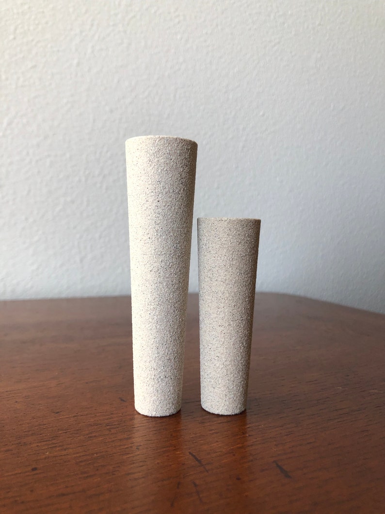 Puede incluir: Dos cilindros de cer&aacute;mica blanca, uno m&aacute;s alto que el otro, con una superficie texturizada. Los cilindros est&aacute;n sobre una superficie de madera.