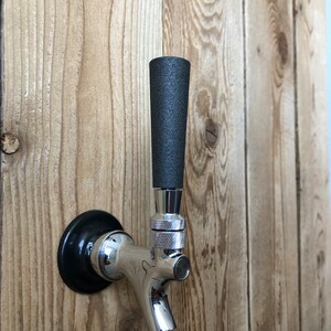 Puede incluir: Un grifo de cerveza cromado con una manija de goma negra montada en una pared de madera.