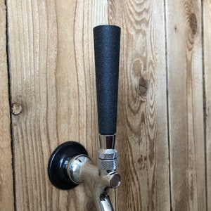 Puede incluir: Un grifo de cerveza negro y plateado con una manija negra. El grifo est&aacute; montado en una superficie de madera.