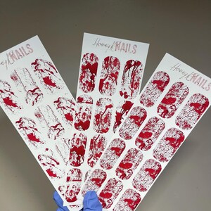 May include: Three white rectangular sheets of nail wraps featuring a red blood splatter pattern. Each sheet displays the text Honey NAILS at the top. The designs include various shapes and sizes of red paint-like drips and speckles on a white background.