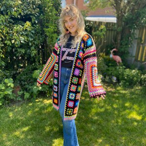 Puede incluir: Una mujer lleva una chaqueta de crochet negra con cuadrados coloridos y una camiseta negra con el texto "Apocalypse Dreams". Lleva pantalones vaqueros azules y botas negras.