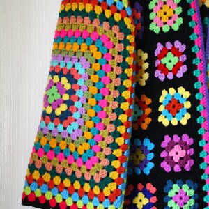 Puede incluir: Una chaqueta de ganchillo colorida con fondo negro y un patr&oacute;n de cuadrado de la abuela arco&iacute;ris en la manga. La chaqueta est&aacute; hecha con una variedad de colores, incluyendo rojo, naranja, amarillo, verde, azul, morado y rosa.
