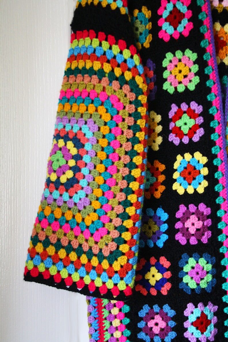 Puede incluir: Una chaqueta de ganchillo colorida con fondo negro y un patr&oacute;n de cuadrado de la abuela arco&iacute;ris en la manga. La chaqueta est&aacute; hecha con una variedad de colores, incluyendo rojo, naranja, amarillo, verde, azul, morado y rosa.