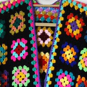 Puede incluir: Una chaqueta de punto de ganchillo colorida con fondo negro y cuadrados de colores arco&iacute;ris. La chaqueta est&aacute; abierta por delante y tiene cuello en V.