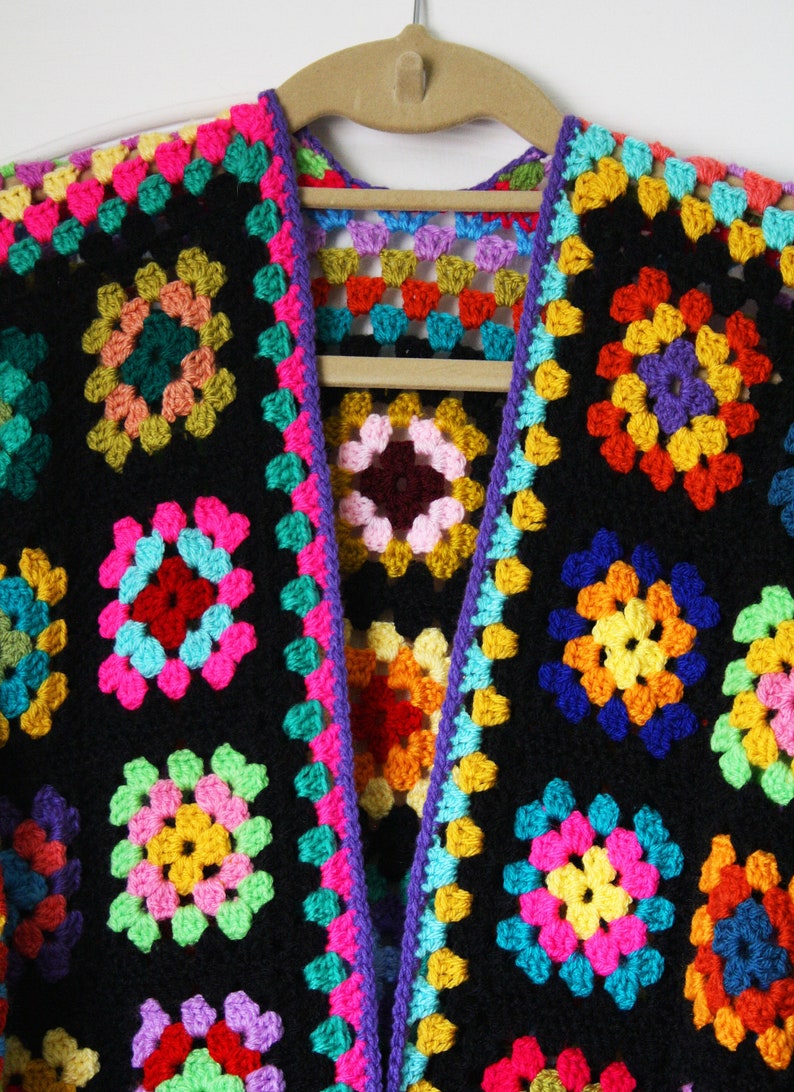Puede incluir: Una chaqueta de punto de ganchillo colorida con fondo negro y cuadrados de colores arco&iacute;ris. La chaqueta est&aacute; abierta por delante y tiene cuello en V.