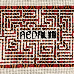 May include: A cross-stitch pattern of a maze with the word "REDRUM" spelled backwards in the center. The maze is surrounded by a red and orange border.