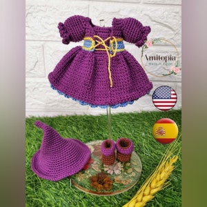 May include: A handmade crochet doll outfit in shades of purple, blue, and yellow. The set includes a dress, hat, and boots. The dress features a bodice with yellow lacing and a flared skirt. The outfit is displayed on a clear stand.