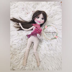 May include: A handmade crochet doll with long brown hair and a delicate face, holding a bouquet of pink flowers. The doll is lying on a fluffy white surface. The doll's skin is a light beige color. The doll is wearing no clothes. The logo "Amitopia Made in Egypt" is visible.