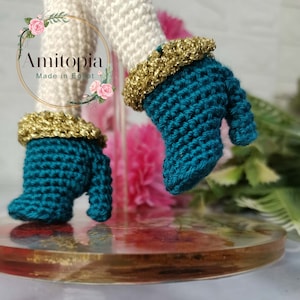 May include: Close-up of teal crochet boots with gold trim, part of a doll or figurine. The boots are on a clear stand with a floral design. The text "Amitopia Made in Egypt" is visible in a circular logo.