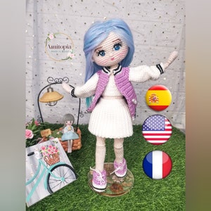 May include: A handmade crochet doll with blue hair, wearing a white dress, a purple vest, and pink sneakers. The doll has blue eyes and is standing on a clear stand. The doll is part of a collection of handmade dolls.