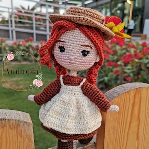 Puede incluir: Muñeca de ganchillo hecha a mano, con cabello rojo, que lleva un sombrero marrón con una flor, un delantal blanco y un vestido marrón. La muñeca está sobre una valla de madera. Un objeto decorativo encantador.