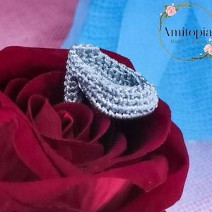 Peut inclure: Gros plan d'un chausson de bébé crocheté gris clair, posé sur une rose rouge foncé. La chaussure est faite à la main avec des détails de points visibles. Le fond présente un tissu rose et bleu doux.