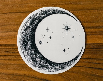May include: A white sticker with a black crescent moon and stars design. The sticker is on a wooden surface.