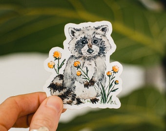 May include: A watercolor sticker featuring a cute raccoon with black and gray fur, sitting amongst yellow flowers. The raccoon has a black nose and black eyes.